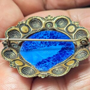 May include: Vintage oval brooch with a blue and white glass center. The brooch has a decorative gold-toned metal frame with intricate detailing. The pin is visible on the back, indicating it can be attached to clothing or accessories.