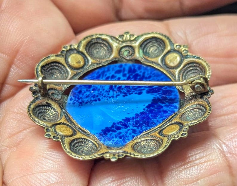 May include: Vintage oval brooch with a blue and white glass center. The brooch has a decorative gold-toned metal frame with intricate detailing. The pin is visible on the back, indicating it can be attached to clothing or accessories.