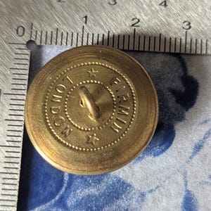 May include: A vintage brass button with the text "LONDON FIRMIN" around the edge. The button has a loop on the back for sewing and is approximately 1 inch in diameter. The button is on a blue and white floral patterned surface.