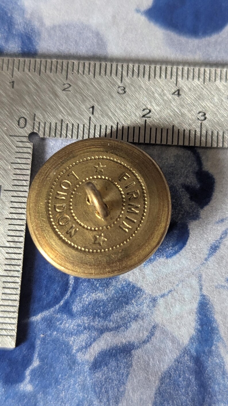 May include: A vintage brass button with the text "LONDON FIRMIN" around the edge. The button has a loop on the back for sewing and is approximately 1 inch in diameter. The button is on a blue and white floral patterned surface.