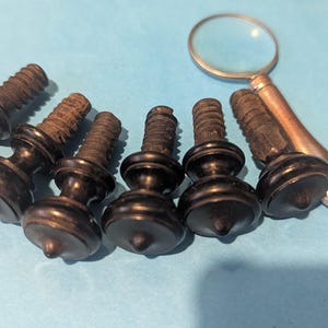 May include: A set of six dark brown, turned wooden furniture knobs with threaded metal screws extending from the back. Each knob features a rounded, tiered profile with a pointed center tip. A small handheld magnifying glass is positioned nearby.