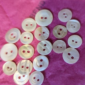 May include: A collection of seventeen round, off-white buttons of varying sizes, each featuring two central holes. These buttons have a smooth, slightly iridescent surface texture and are arranged in a scattered pattern on a bright pink background.