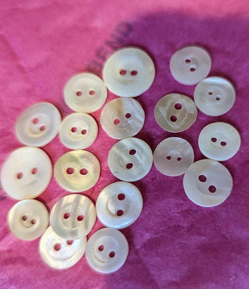 May include: A collection of seventeen round, off-white buttons of varying sizes, each featuring two central holes. These buttons have a smooth, slightly iridescent surface texture and are arranged in a scattered pattern on a bright pink background.