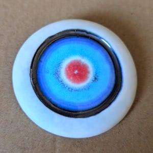 Puede incluir: Un objeto cerámico redondo con un borde exterior blanco, un anillo interior marrón oscuro y un diseño de diana azul y rojo en el centro. El objeto tiene un acabado brillante y una forma ligeramente irregular. El diseño se asemeja a una diana.
