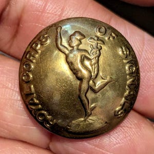 Op de afbeelding: Een ronde, messingkleurige metalen knoop met een reliëf van Mercurius die een caduceus vasthoudt. De tekst ROYAL CORPS OF SIGNALS is langs de buitenrand van de knoop in reliëf aangebracht en omcirkelt de centrale figuur in een militair ontwerp.