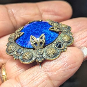 May include: Vintage oval brooch with a vibrant blue stone center. The brooch features an ornate, antique gold-tone metal frame with leaf and floral details, and small black accents. A classic piece of jewelry.