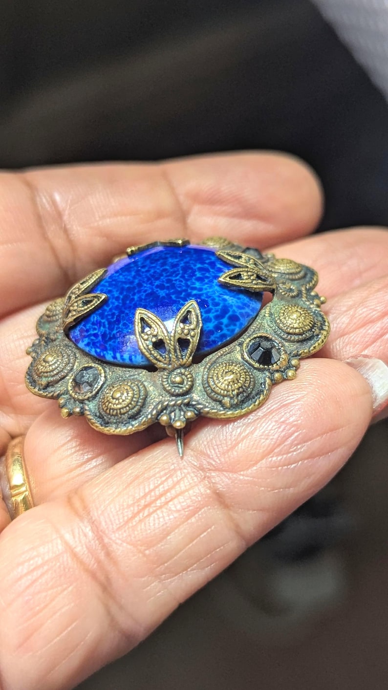 May include: Vintage oval brooch with a vibrant blue stone center. The brooch features an ornate, antique gold-tone metal frame with leaf and floral details, and small black accents. A classic piece of jewelry.