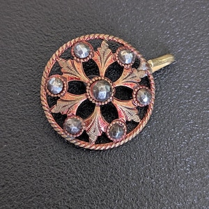 May include: A round, antique brooch with a floral design. The brooch features a central flower shape surrounded by eight smaller, gem-like stones. The metal has a copper-like color with a twisted rope border and a gold-colored pin.