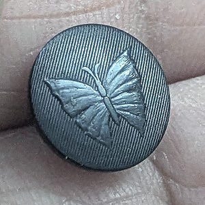 May include: A black button with a raised, detailed butterfly design. The button has a textured surface.