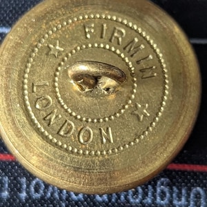 May include: Close-up of a vintage brass button with the text "FIRMIN LONDON" encircling the center. The button has a loop on the back for sewing and is set against a dark background.
