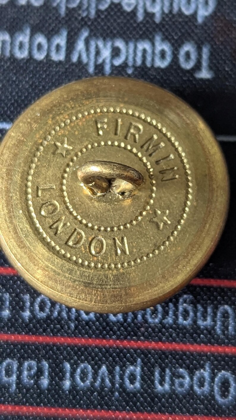 May include: Close-up of a vintage brass button with the text "FIRMIN LONDON" encircling the center. The button has a loop on the back for sewing and is set against a dark background.