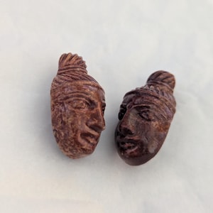 May include: Two small, carved sculptures of human faces. The faces are brown and have detailed features, including eyes, noses, and mouths. The hair is styled in a bun. The sculptures are on a white surface.