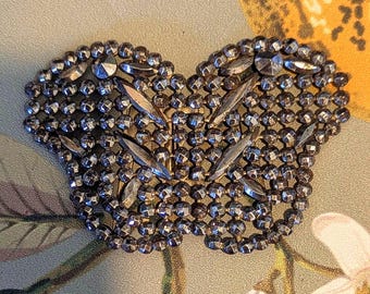 May include: A vintage silver-toned metal brooch with a heart-shaped design. The brooch is encrusted with small, faceted beads and larger, elongated crystal accents. The background features a floral pattern.
