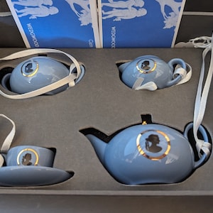 May include: A Wedgwood blue tea set in a black box. The set includes a teapot, creamer, sugar bowl, and a teacup and saucer. The tea set is decorated with a gold trim and a silhouette of a woman.