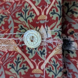 May include: A fabric cuff featuring a repeating botanical pattern in shades of deep red, sage green, and cream. The cuff is secured with a single, round, iridescent white button with a textured, radial grooved design and visible stitching.
