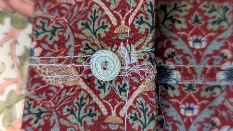 May include: A fabric cuff featuring a repeating botanical pattern in shades of deep red, sage green, and cream. The cuff is secured with a single, round, iridescent white button with a textured, radial grooved design and visible stitching.