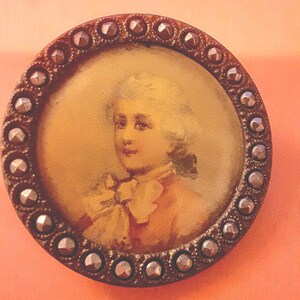 May include: A round, antique-looking button with a silver-coloured metal rim and a raised, textured design. The centre of the button features a portrait of a person with a white wig and a white collar, painted on a yellow background.