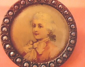 May include: A round, antique-looking button with a silver-colored metal rim and a raised, textured design. The center of the button features a portrait of a person with a white wig and a white collar, painted on a yellow background.