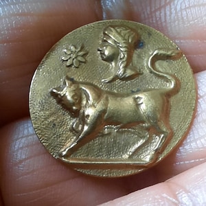 May include: A gold-toned metal button with a raised design of a bull and a woman's head. The bull is facing right and the woman's head is facing left. There is a small flower design above the woman's head.