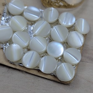 May include: A collection of fifteen round, white, iridescent buttons arranged in a grid on a rectangular backing, accompanied by a gold-toned metal ring featuring an intricate, cut-out filigree pattern along the top portion of the band.