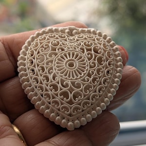 May include: A heart-shaped off-white brooch featuring an intricate filigree pattern with a central circular sunburst motif and a beaded border. The design consists of delicate, raised scrollwork and symmetrical curves that create a lace-like texture.