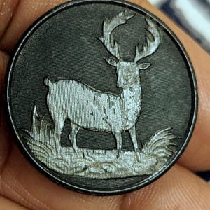 Op de afbeelding: Een ronde, donkergrijze metalen knoop met een gedetailleerd reliëf van een hert dat in hoog gras staat. Het hert is lichtgrijs met opvallende geweien. De knoop heeft een getextureerde rand en een licht versleten uiterlijk.