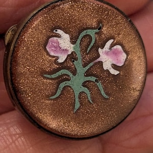 Op de afbeelding: Een ronde, antieke sieradendoos met een glinsterend, bronskleurig oppervlak. De deksel heeft een bloemenontwerp met roze en witte bloemen en groene stelen. De doos heeft een donkerbruine rand.
