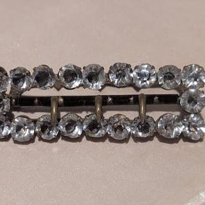 May include: A rectangular metal buckle featuring a frame lined with clear, faceted rhinestones. The buckle has a dark, metallic finish on the central bar and three small, curved prongs extending from the bottom edge of the frame.