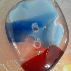 May include: A clear, irregularly shaped glass object with embedded colors. The glass features sections of light blue, dark blue, and red. It has two holes and several small bubbles within the glass, creating a unique decorative piece.