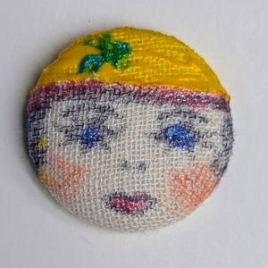Könnte beinhalten: Ein runder Stoffknopf mit einem gemalten Gesicht. Der Knopf hat eine gelbe Kopfbedeckung mit grünem Akzent, blaue Augen, rosa Wangen und einen rosa Mund. Das Gesicht ist auf einem weißen Stoff gestickt.