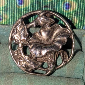 Op de afbeelding: Zilverkleurige, ronde broche met een bloemmotief, inclusief bladeren en een grote, getextureerde bloem. De broche heeft een open ontwerp en ligt op een turquoise achtergrond.