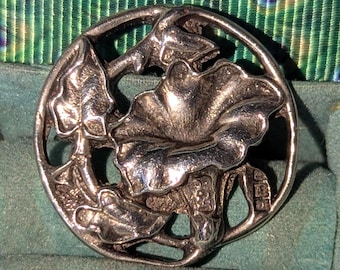 May include: A silver-toned, circular brooch featuring a floral design with leaves and a large, textured flower. The brooch has an openwork design and is set against a teal background.