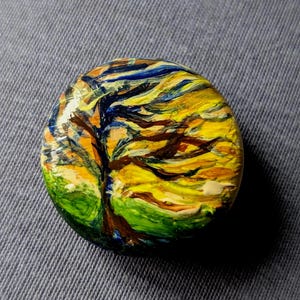 Op de afbeelding: Een rond, handgeschilderd kunstwerk met een boom met een levendige gele en oranje lucht. De takken van de boom zijn donkerbruin tegen de kleurrijke achtergrond. Groen gras staat onderaan. Het kunstwerk staat op een grijze ondergrond.