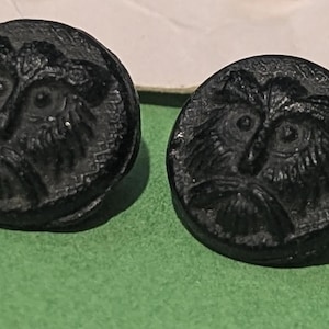 May include: Two round black buttons featuring a moulded relief design of a stylised owl face with prominent eyes, a beak, and textured feather details. The buttons have a circular shape and a dark, matte finish, showcasing intricate craftsmanship.