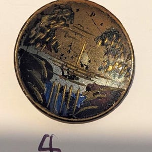 May include: A circular metal button featuring a weathered landscape illustration with gold, black, and blue tones. The design depicts stylized trees and a bridge over water, showing significant surface wear and patina across the metallic finish.