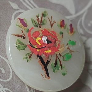 Op de afbeelding: Een ronde, crèmekleurige knoop met een handgeschilderd bloemmotief. De centrale bloem is rood met gele accenten, omgeven door groene bladeren en bruine stelen. De knoop heeft een diameter van ongeveer 3,8 cm.
