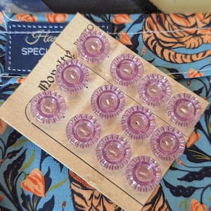 Pode incluir: Um conjunto de doze botões lilás claros com borda ondulada em um suporte de papelão. Os botões são dispostos em um tecido com estampa floral. A etiqueta diz "Handmade Speciality".