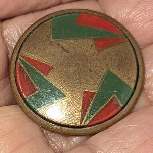 Op de afbeelding: Een ronde knoop in antieke stijl met een bronzen metalen frame. Het midden heeft een geometrisch ontwerp van rode en groene vormen op een getextureerde, bronzen achtergrond. De knoop wordt in een hand gehouden.