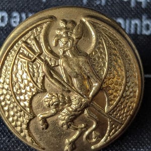 May include: A gold-colored button with a detailed relief of a winged creature holding a trident. The button has a circular shape and is likely made of metal. The design is intricate and well-defined.