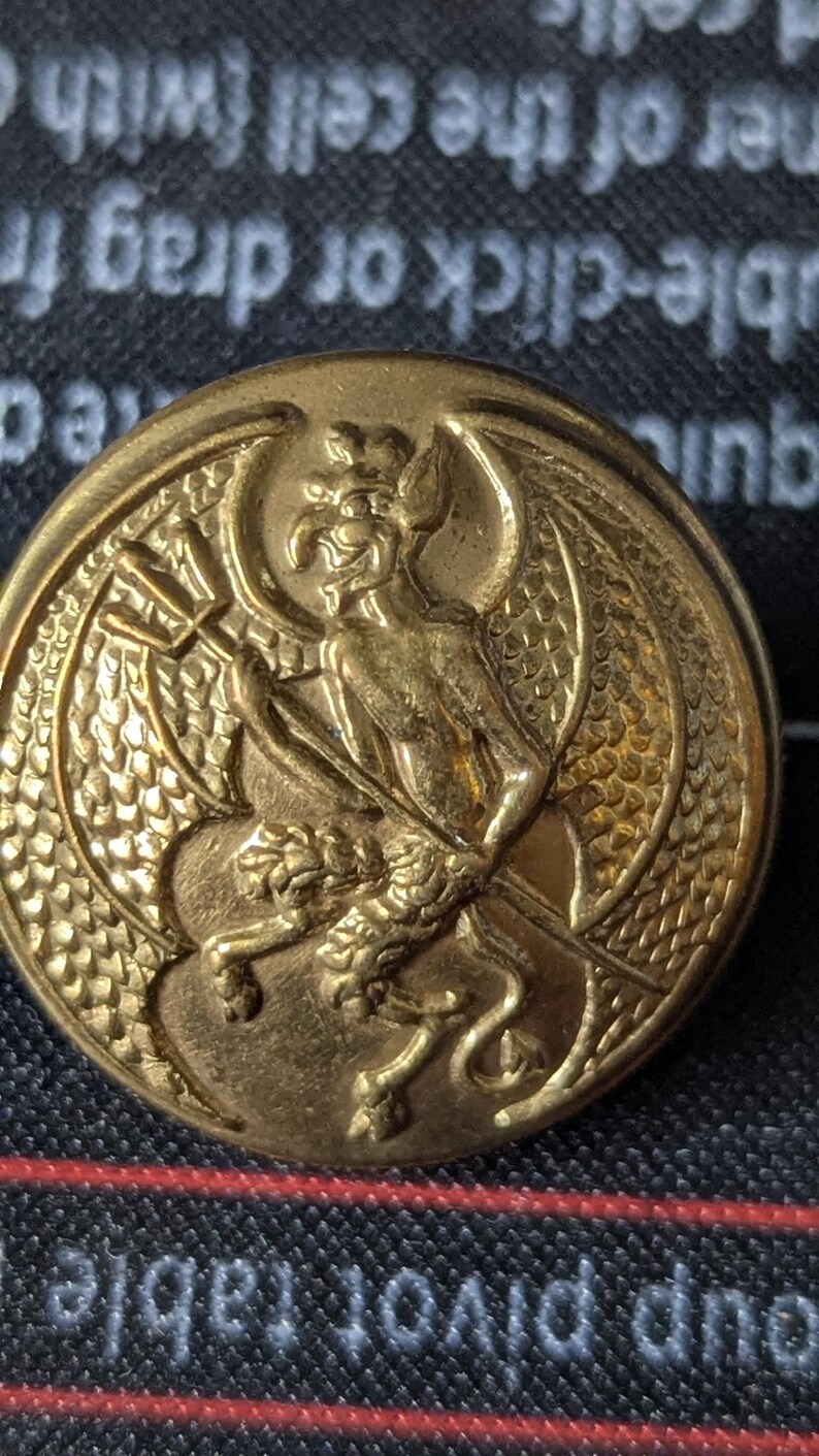 May include: A gold-colored button with a detailed relief of a winged creature holding a trident. The button has a circular shape and is likely made of metal. The design is intricate and well-defined.