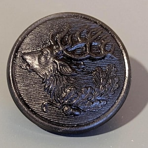 Op de afbeelding: Een donkergrijze knoop met een gedetailleerd reliëf van een hertenkop, gewei en bladeren. De knoop is rond met een getextureerd oppervlak en een opstaande rand. Een klassiek ontwerp voor kleding.