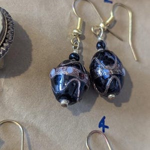 Peut inclure: Une paire de boucles d'oreilles pendantes composées de perles de verre ovales noires avec des motifs tourbillonnants en cuivre métallique et de petits accents blancs. Les perles sont suspendues à des crochets en métal doré avec des perles noires.