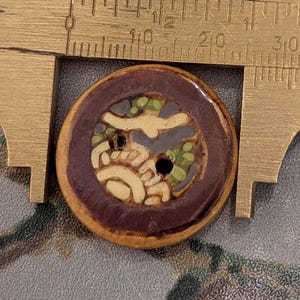 Op de afbeelding: Een ronde, decoratieve knoop met een bruine rand en een centraal ontwerp met crème, groene en grijze elementen. De knoop heeft twee gaten om te naaien en wordt gemeten met een messing schuifmaat.