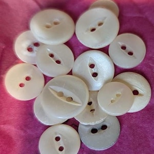 May include: A collection of round, off-white buttons with a pearlescent finish. Each button features two central holes, some with a recessed channel connecting the holes. The buttons vary slightly in size and are scattered across a pink surface.