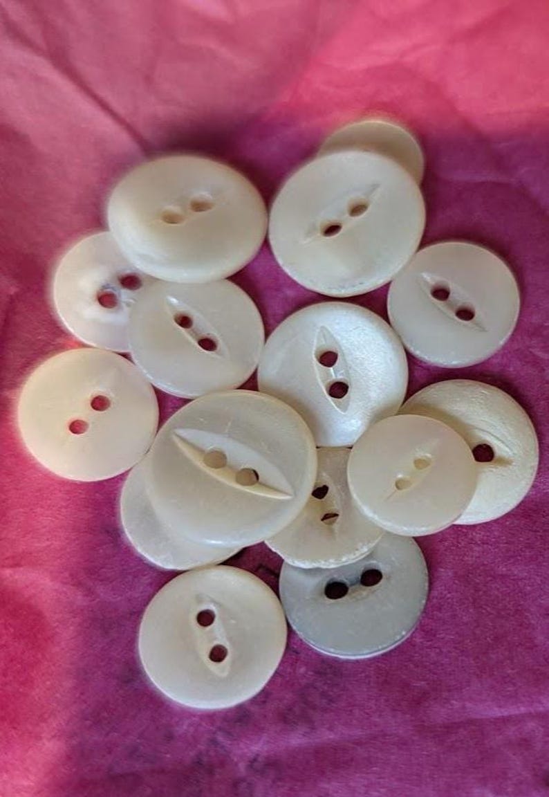 May include: A collection of round, off-white buttons with a pearlescent finish. Each button features two central holes, some with a recessed channel connecting the holes. The buttons vary slightly in size and are scattered across a pink surface.