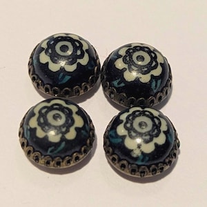 Puede incluir: Cuatro botones redondos vintage. Cada botón presenta un fondo azul oscuro con un diseño floral blanco y hojas turquesas. Tienen un borde decorativo de metal color bronce.