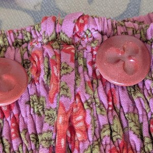 Op de afbeelding: Een roze kledingstuk met een geplooide, gesmokte textuur en een bloemenpatroon in rood- en groentinten. Twee ronde, roodbruine knopen met een drielobbige, klaverachtige inkeping zijn aan de voorzijde van de stof bevestigd.