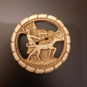 Op de afbeelding: Een ronde, beige knoop met een gebeeldhouwde jachtscène. Het ontwerp toont een persoon met een hond, een gebouw en een decoratieve rand. De knoop heeft twee gaten om te naaien.