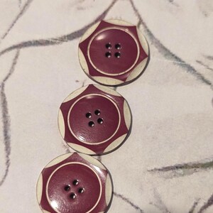 Op de afbeelding: Drie decoratieve knopen met een bordeauxrood midden en een crèmekleurige, stervormige rand. Elke knoop heeft vier gaten om te naaien. De knopen zijn gerangschikt op een patroon, gebroken witte ondergrond.