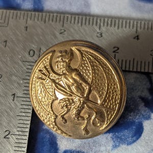 May include: A gold-colored button with a detailed embossed design of a devil figure holding a trident. The devil has wings and is seated on a horned creature. The button is round and has a diameter of approximately 1 inch.